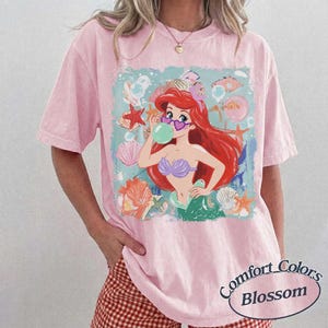 May include: A light pink short-sleeve t-shirt featuring a graphic of a red-haired mermaid wearing heart-shaped sunglasses and blowing a bubble. The design includes various seashells and starfish in shades of orange, pink, and white on a light blue background.