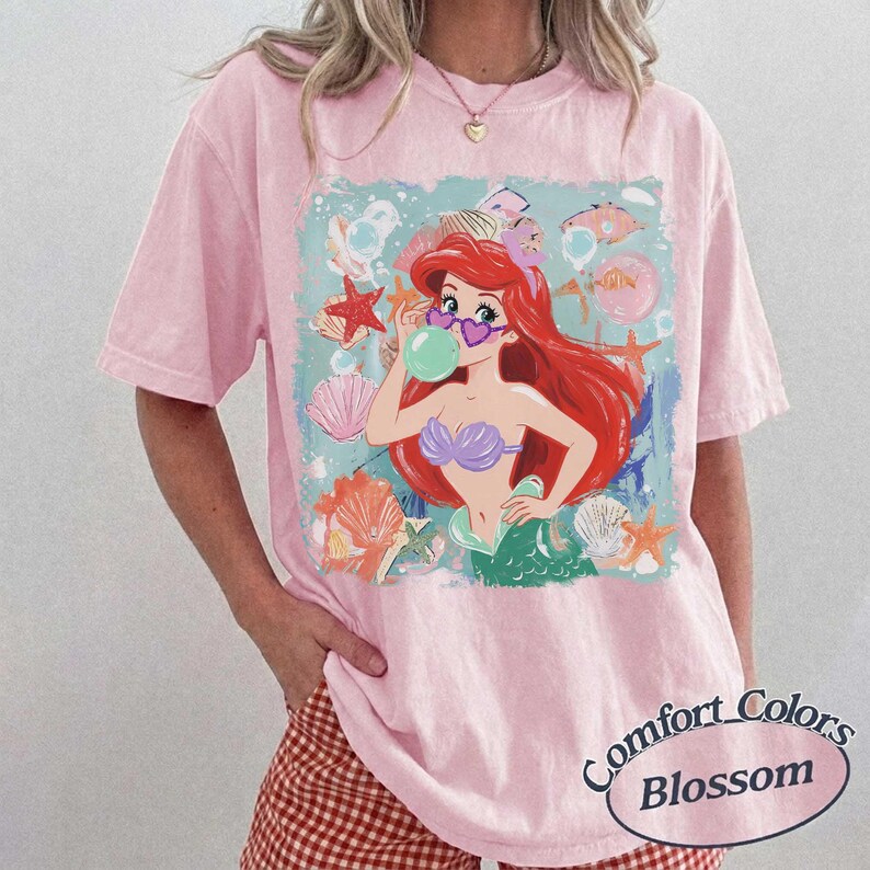 May include: A light pink short-sleeve t-shirt featuring a graphic of a red-haired mermaid wearing heart-shaped sunglasses and blowing a bubble. The design includes various seashells and starfish in shades of orange, pink, and white on a light blue background.