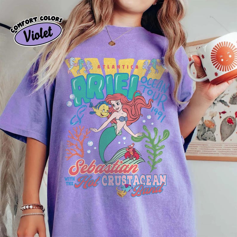 May include: A violet short-sleeve t-shirt featuring a graphic print of Ariel, Flounder, and Sebastian from The Little Mermaid. The text reads Atlantica Ocean Tour 1991, Sebastian with the Hot Crustacean Band, and includes coral and seaweed illustrations.