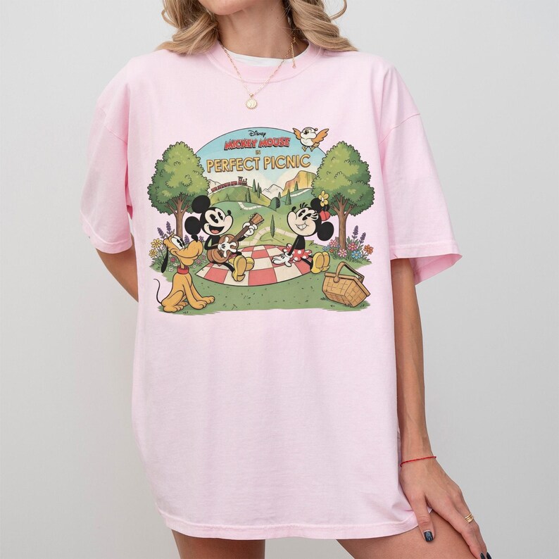 May include: Light pink t-shirt featuring a cartoon scene with Mickey Mouse, Minnie Mouse, and Pluto having a picnic. The text "Disney PERFECT PICNIC" is arched above the scene. The shirt has short sleeves and a relaxed fit.