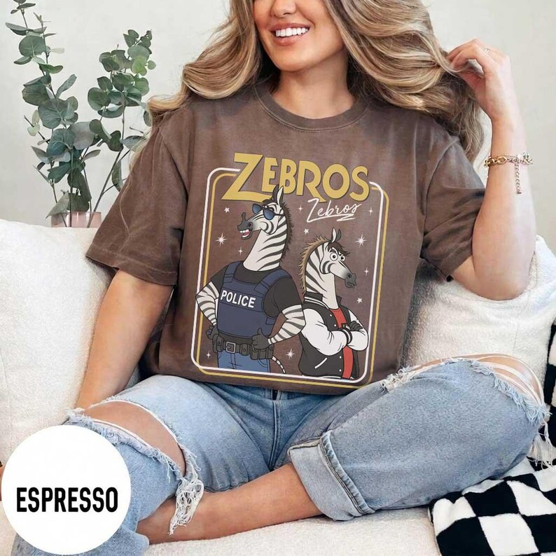 May include: Espresso-coloured t-shirt with a cartoon graphic of two zebras. One zebra wears a police vest, the other a jacket. The text "ZEBROS" is above the zebras. The shirt has a relaxed fit.