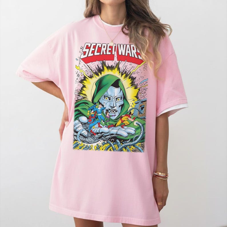 May include: Pink t-shirt with a comic book graphic featuring Doctor Doom and the words "SECRET WARS" in red and silver. The shirt has a white collar and short sleeves.