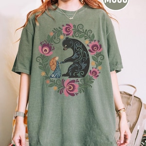 May include: Moss green t-shirt featuring a black bear and a woman in a floral wreath design. The bear has decorative patterns. The woman is wearing a blue dress. The shirt has short sleeves and a crew neck.