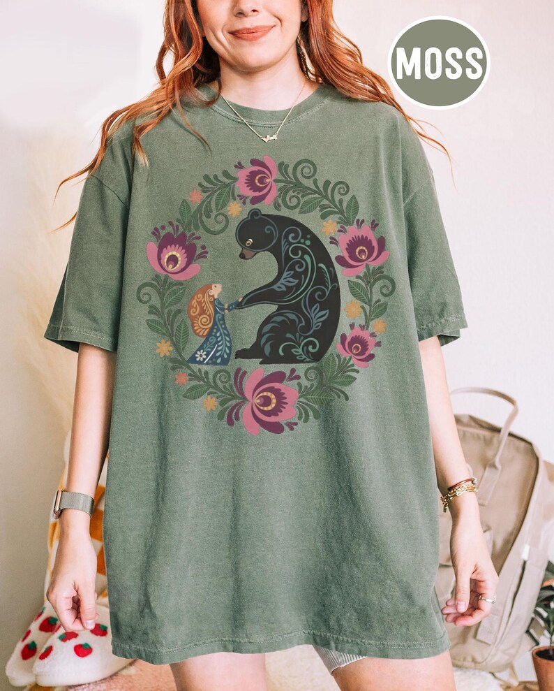 May include: Moss green t-shirt featuring a black bear and a woman in a floral wreath design. The bear has decorative patterns. The woman is wearing a blue dress. The shirt has short sleeves and a crew neck.