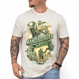 May include: A cream-colored short-sleeve t-shirt featuring a graphic print of The Mandalorian and Grogu. The design includes the text THE MANDALORIAN AND GROGU and THIS IS THE WAY, with illustrations of characters and vehicles from the Star Wars series.