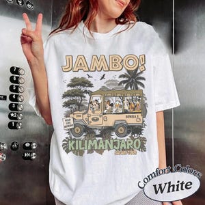 May include: White t-shirt with a safari-themed graphic. The design features a tan jeep with cartoon characters and the text "JAMBO!" and "KILIMANJARO SAFARI TOUR". The shirt also has the text "WILD LIFE RESERVE" and "SIMBA 1".