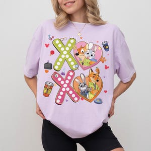 May include: A light purple t-shirt featuring the characters from Zootopia. The shirt has two large "X" shapes, one green and one pink, with cartoon images of Nick Wilde and Judy Hopps inside hearts. Additional cartoon elements are scattered around the design.