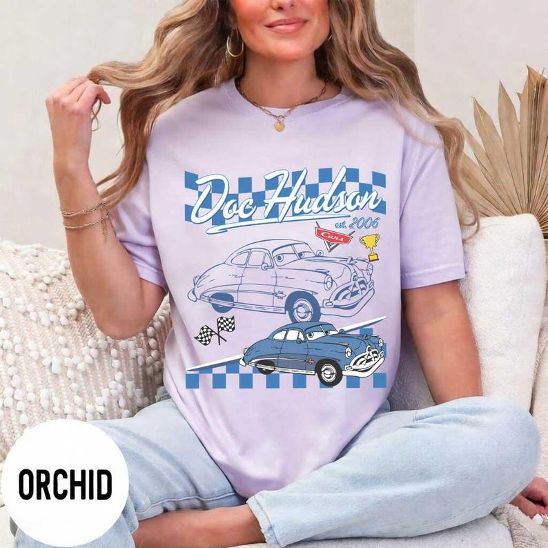 May include: A lilac t-shirt with a graphic of two vintage cars and the text "Doc Hudson est. 2006" in a blue chequered design. The shirt has short sleeves and a crew neck.