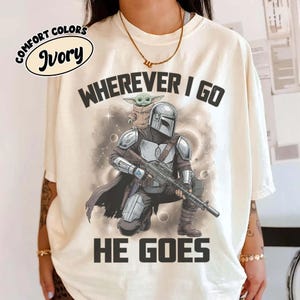 Puede incluir: Una camiseta de color marfil con un gr&aacute;fico de un personaje mandaloriano con armadura plateada arrodillado mientras sostiene un rifle, con una peque&ntilde;a criatura verde sobre el hombro. El texto "WHEREVER I GO HE GOES" est&aacute; impreso en negro.
