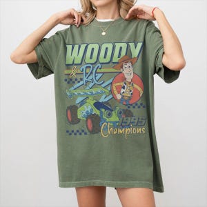 May include: Olive green t-shirt featuring a graphic of Woody and RC from Toy Story. The design includes the text "WOODY & RC 1995 Champions" in green, yellow, and blue. The shirt has short sleeves and a relaxed fit.