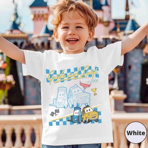 May include: White t-shirt with the text "Guido and Luigi est. 2006" and cartoon car illustrations. The design incorporates a blue and white chequered pattern and a gold trophy. The shirt is worn by a child.