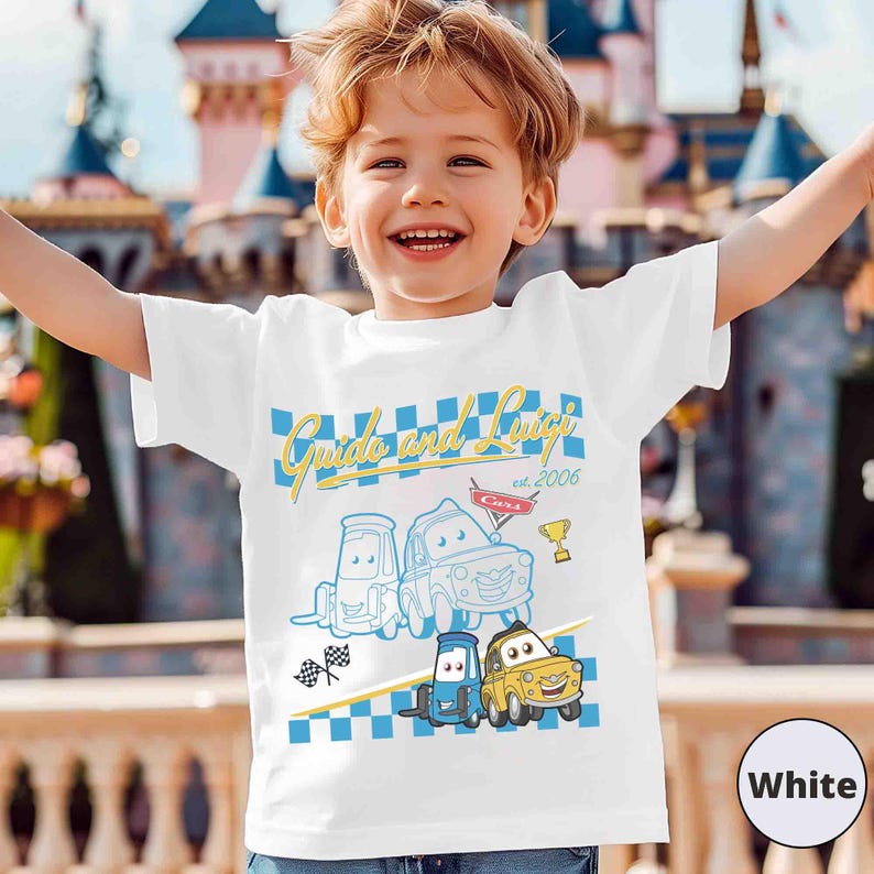 May include: White t-shirt with the text "Guido and Luigi est. 2006" and cartoon car illustrations. The design incorporates a blue and white chequered pattern and a gold trophy. The shirt is worn by a child.