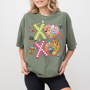 May include: An olive green t-shirt featuring a colorful graphic design. The design includes the letters "X" and "O" made of polka dots, and cartoon characters inside heart shapes. The shirt has short sleeves and a relaxed fit.