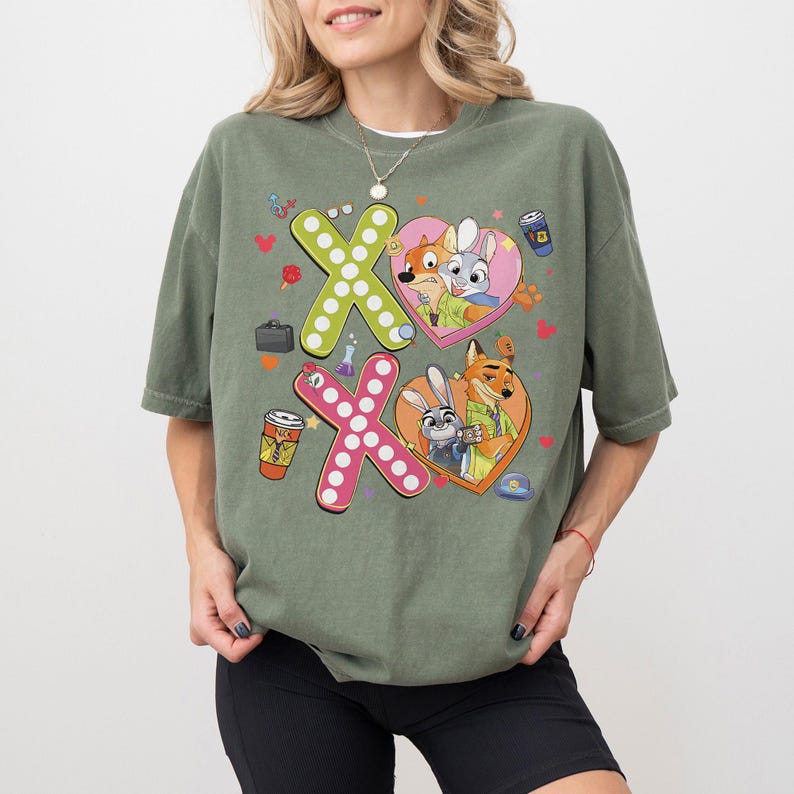 May include: An olive green t-shirt featuring a colorful graphic design. The design includes the letters "X" and "O" made of polka dots, and cartoon characters inside heart shapes. The shirt has short sleeves and a relaxed fit.