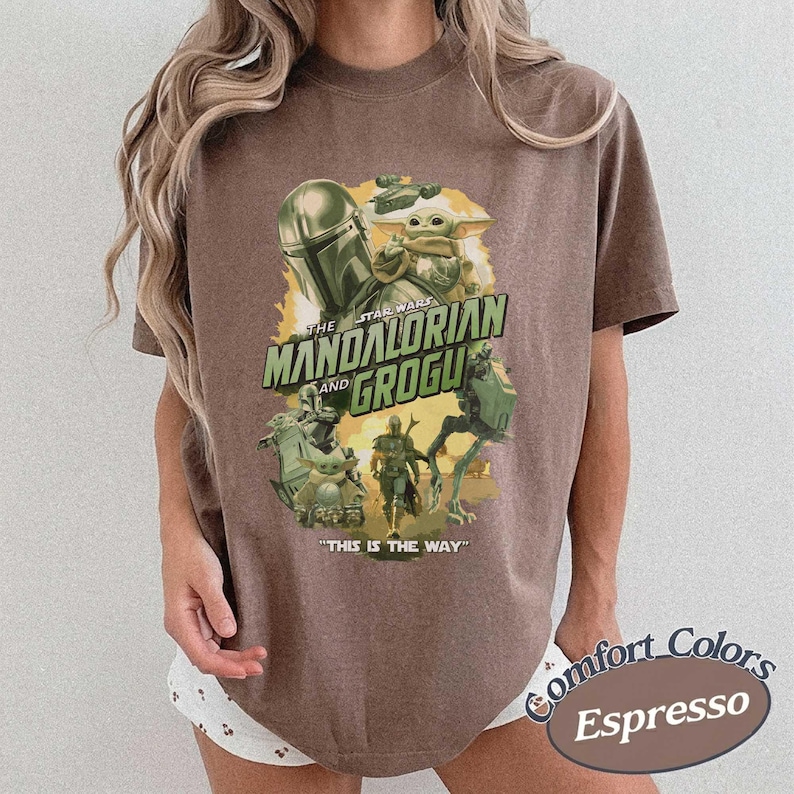 May include: A brown short-sleeve t-shirt featuring a graphic print of The Mandalorian and Grogu. The design includes the text "The Mandalorian and Grogu" and "This is the way" in green, with illustrations of characters and vehicles on a yellow background.