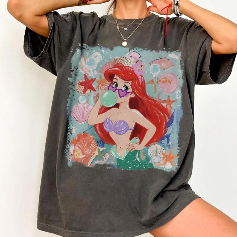 May include: A charcoal gray short-sleeve t-shirt featuring a graphic print of a red-haired mermaid wearing heart-shaped sunglasses and blowing a green bubble. The design includes various seashells and starfish in a light blue, watery background.