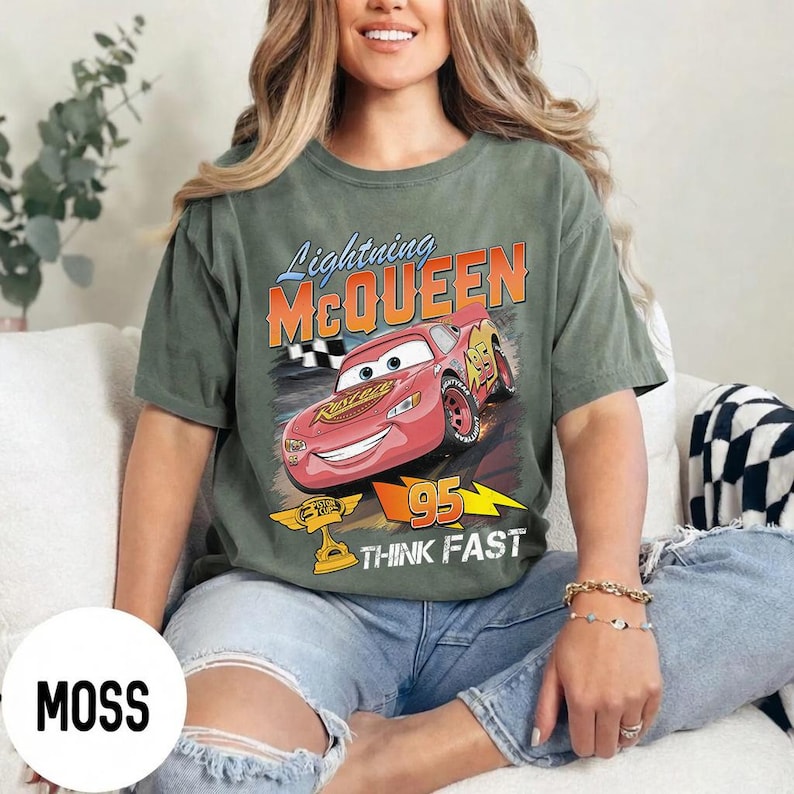 May include: Moss green t-shirt featuring a graphic of Lightning McQueen from the movie Cars. The design includes the text "Lightning McQueen", "95", and "Think Fast". The shirt is a relaxed fit.