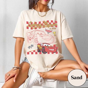 May include: Sand-coloured t-shirt featuring a graphic of Lightning McQueen from the movie Cars. The design includes the text "Lightning McQueen est. 2006" and a red and white checkered pattern. The shirt has short sleeves.