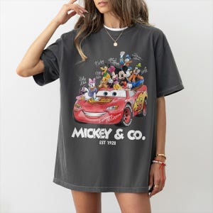May include: Dark gray t-shirt featuring a colorful graphic of Disney characters, including Mickey Mouse, on a red race car. The text "Mickey & Co. EST 1928" is printed below the car. A fun, casual tee.