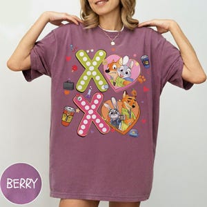 May include: Berry-colored t-shirt featuring the characters from Zootopia. The shirt has the text "XO" with cartoon images of the characters inside hearts. The shirt also has other cartoon images like a coffee cup and a briefcase.