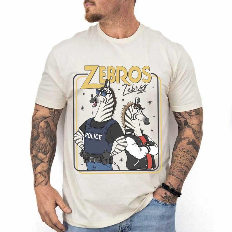 May include: Cream-coloured t-shirt with a cartoon graphic of two zebras. One zebra is dressed as a police officer, and the other wears a jacket. The text "ZEBROS" is displayed above the zebras. The shirt is made of a soft material.