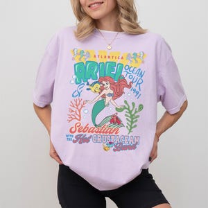 May include: A light purple t-shirt featuring a graphic print of Ariel from The Little Mermaid with the text Atlantica Ocean Tour 1991, Sebastian with the Hot Crustacean Band, and illustrations of Flounder, coral, and seaweed in blue, red, and green.