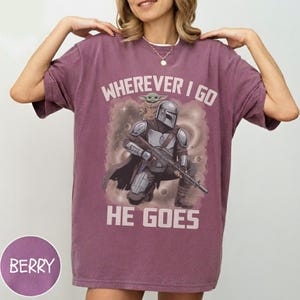 Puede incluir: Una camiseta de manga corta color baya con un gr&aacute;fico de un personaje con casco y armadura que sostiene un rifle, con una peque&ntilde;a criatura verde sobre el hombro. El texto "WHEREVER I GO HE GOES" est&aacute; impreso en blanco sobre y bajo la ilustraci&oacute;n.