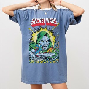 May include: A blue t-shirt featuring a comic book graphic. The graphic includes the text "SECRET WARS" in red and white, along with a depiction of a character in a green hood and metallic mask, and other comic book characters.