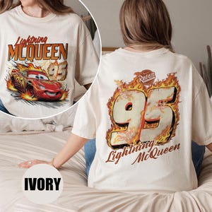 May include: Ivory-colored t-shirt featuring a graphic of Lightning McQueen from the Cars movie. The front design shows the car with flames and the text "Lightning McQueen 95." The back displays "95" in flames and the text "Lightning McQueen."