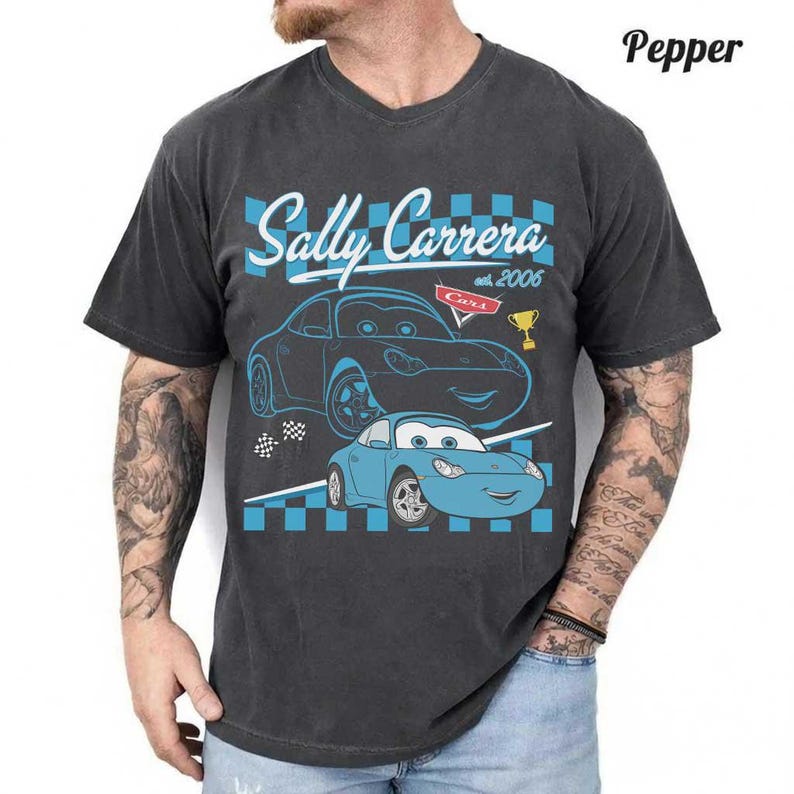 May include: Dark grey t-shirt with a graphic of Sally Carrera from the film Cars. The design includes the text "Sally Carrera" and "Cars est. 2006" with a blue and white chequered pattern.