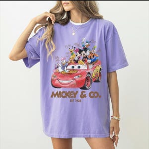 May include: A lilac-colored t-shirt featuring a graphic of Disney characters riding in a red race car. The car has the number 95 on it. The text "Mickey & Co. Est 1928" is printed below the car.