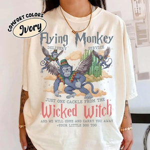 May include: Ivory-colored t-shirt featuring a graphic of a flying monkey with wings, a green hat, and a red and green collar. The text reads "Flying Monkey Delivery Service" and "Just One Cackle From The Wicked Witch".