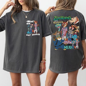 May include: Dark grey t-shirt featuring a colorful graphic from the movie Zootopia. The front of the shirt has the text "Not on our watch" with cartoon characters. The back of the shirt has the text "Zootopia" and "Let's Boogie".