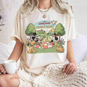 May include: Cream-colored t-shirt featuring a graphic of Mickey Mouse, Minnie Mouse, and Pluto having a picnic. The design includes the text "Mickey Mouse Perfect Picnic" in a vintage cartoon style. The shirt is a relaxed fit.