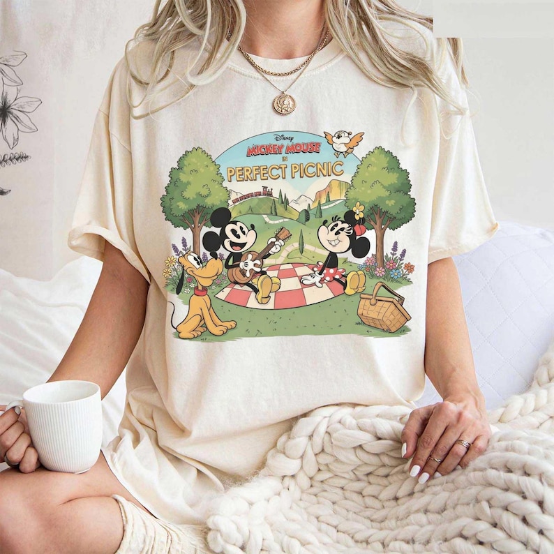 May include: Cream-colored t-shirt featuring a graphic of Mickey Mouse, Minnie Mouse, and Pluto having a picnic. The design includes the text "Mickey Mouse Perfect Picnic" in a vintage cartoon style. The shirt is a relaxed fit.