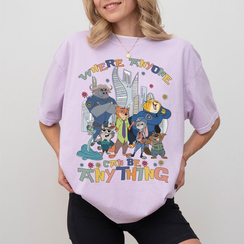 Puede incluir: Camiseta lila claro con un gr&aacute;fico colorido de personajes de dibujos animados y el texto "WHERE ANYONE CAN BE ANYTHING". El dise&ntilde;o incluye un paisaje urbano y varias figuras de animales.