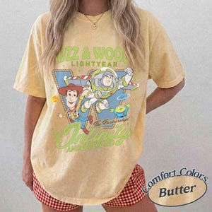 Peut inclure: Un t-shirt jaune beurre &agrave; manches courtes avec une impression graphique de Woody, Buzz l'&Eacute;clair et un extraterrestre de Toy Story. Le texte indique BUZZ & WOODY LIGHTYEAR, The Partnership!, et TO INFINITY AND BEYOND en lettres vertes et blanches.