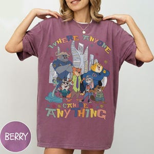 Puede incluir: Camiseta de color bayas con un gr&aacute;fico colorido de personajes de dibujos animados y el texto "WHERE ANYONE CAN BE ANYTHING". El dise&ntilde;o incluye un paisaje urbano y varios personajes de animales con uniformes de polic&iacute;a y ropa informal.