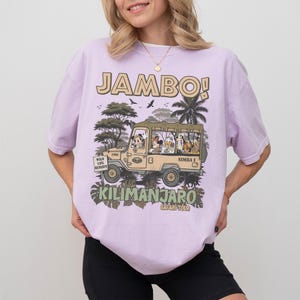 May include: A light purple t-shirt with a safari-themed graphic. The graphic features a tan jeep with cartoon characters and the words "JAMBO!" and "Kilimanjaro Safari Tour". The shirt has short sleeves.