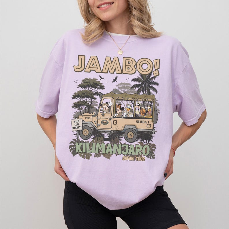 May include: A light purple t-shirt with a safari-themed graphic. The graphic features a tan jeep with cartoon characters and the words "JAMBO!" and "Kilimanjaro Safari Tour". The shirt has short sleeves.