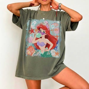May include: An olive green short-sleeve t-shirt featuring a graphic print of a red-haired mermaid wearing heart-shaped sunglasses and blowing a green bubble. The design includes various seashells and starfish in shades of pink, orange, and blue.