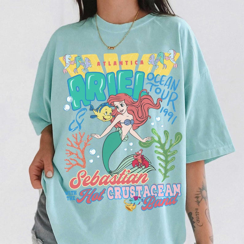 May include: A light blue t-shirt featuring a graphic print of Ariel, Flounder, and Sebastian. The text reads Atlantica Ocean Tour 1991, Ariel, Sebastian, and The Hot Crustacean Band. The design includes colorful coral, seaweed, and bubbles.