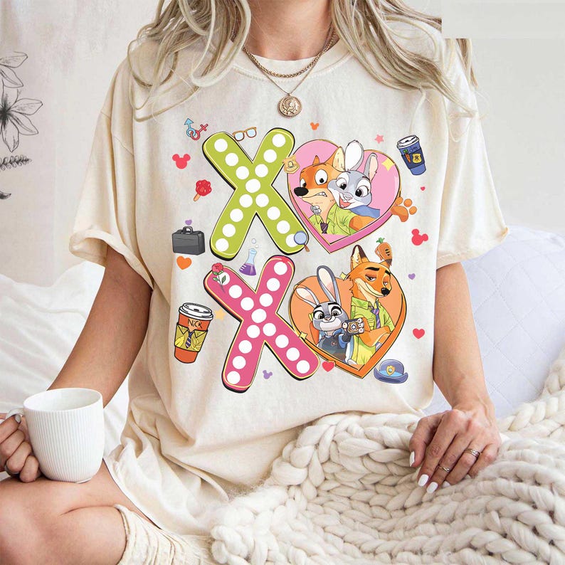 May include: Cream-colored t-shirt featuring a graphic design with the characters from Zootopia. The design includes two "XO" shapes, each with a heart-shaped image of the characters. Additional cartoon elements are scattered around the design.