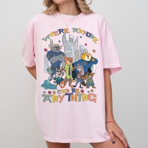 Puede incluir: Camiseta rosa claro con un estampado colorido de personajes de la pel&iacute;cula animada Zootopia. El texto "WHERE ANYONE CAN BE ANYTHING" est&aacute; impreso encima y debajo de los personajes. La camiseta tiene mangas cortas y un corte holgado.