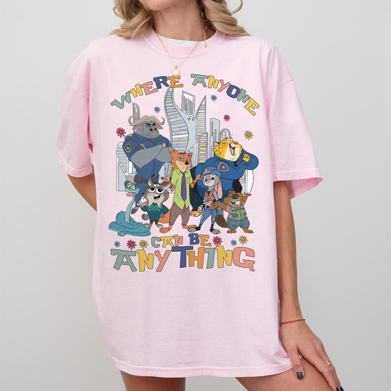 Puede incluir: Camiseta rosa claro con un estampado colorido de personajes de la pel&iacute;cula animada Zootopia. El texto "WHERE ANYONE CAN BE ANYTHING" est&aacute; impreso encima y debajo de los personajes. La camiseta tiene mangas cortas y un corte holgado.