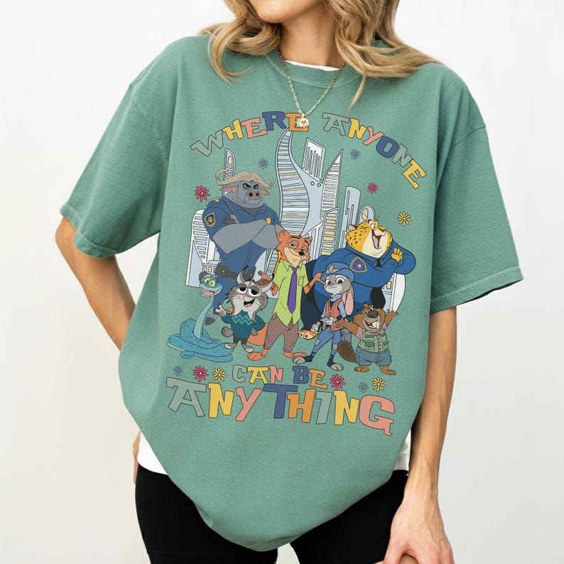 Puede incluir: Camiseta verde con un gr&aacute;fico colorido de personajes de dibujos animados y el texto "WHERE ANYONE CAN BE ANYTHING". El dise&ntilde;o incluye un paisaje urbano y varias figuras de animales con uniformes de polic&iacute;a y ropa informal.