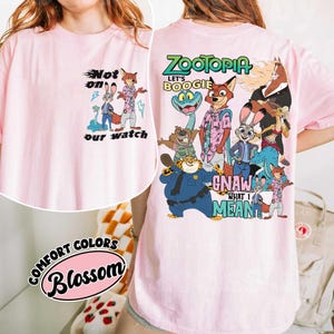 May include: A light pink t-shirt featuring cartoon characters from Zootopia. The front of the shirt has the text "Not on our watch". The back of the shirt has the text "Zootopia Let's Boogie Gnaw What I Mean". The shirt is labeled "Comfort Colors Blossom".