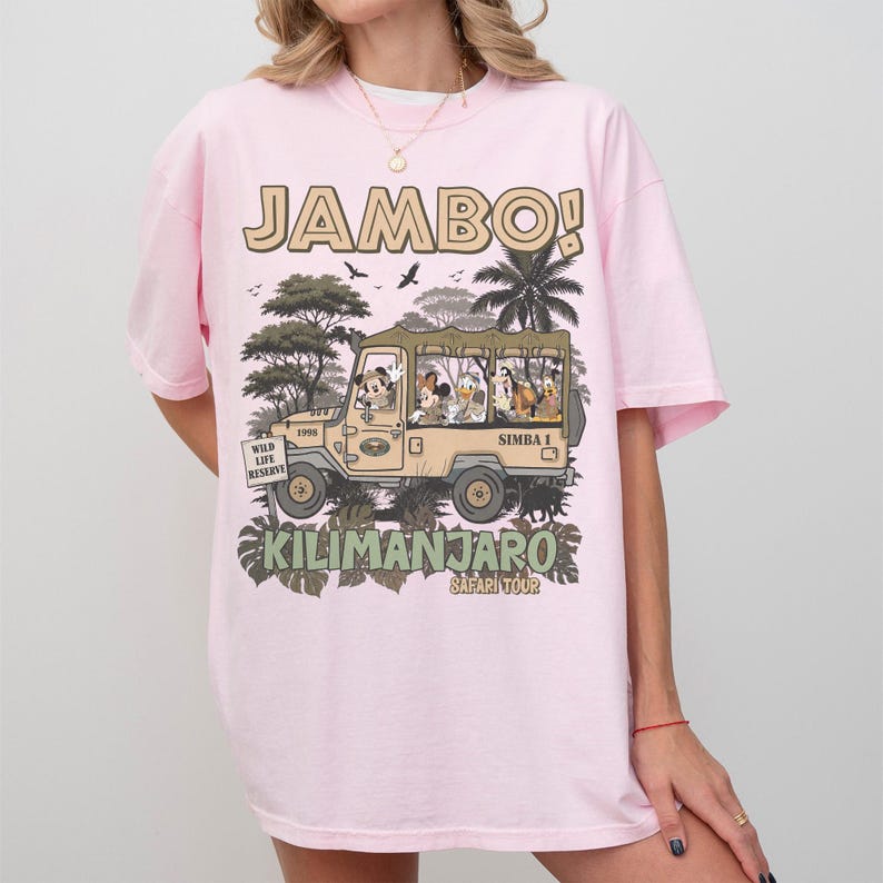 May include: Pink t-shirt featuring a safari scene with Mickey Mouse and friends in a jeep. The shirt has the words "JAMBO!" and "Kilimanjaro Safari Tour" printed on it. The jeep is labeled "Simba 1" and the shirt is a light pink color.