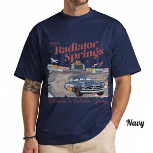 May include: Navy blue t-shirt featuring a graphic of a blue car racing on a track with the words "Radiator Springs" and "Welcome to Radiator Springs" in red. The shirt also has the text "California est. 2012".