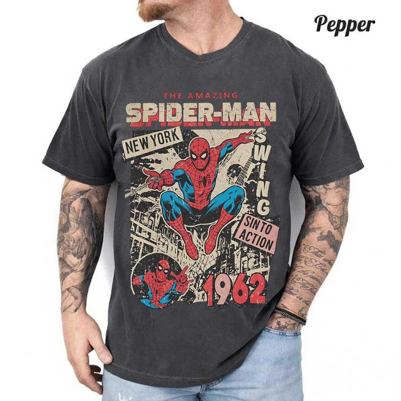 Pode incluir: T-shirt cinzenta escura com um gr&aacute;fico do Homem-Aranha. O design inclui as palavras "The Amazing Spider-Man", "New York" e o ano "1962". A ilustra&ccedil;&atilde;o mostra o Homem-Aranha no seu fato vermelho e azul.